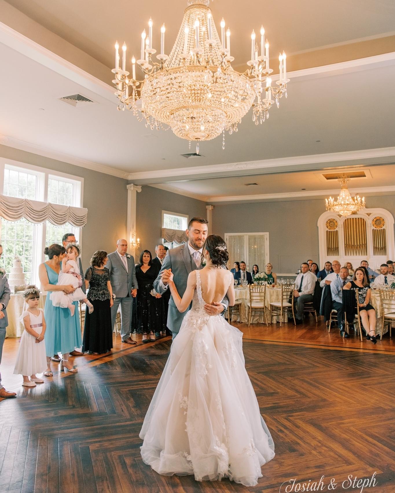 Wedding couple first dance under grand chandelier and guest watching.